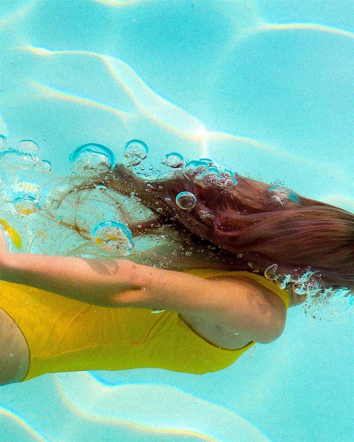 Ung kvinne som svømmer under vann (kun overkroppen og det lange håret hennes er synlig, ansiktet vises ikke). Hun har gul badedrakt. - Klikk for stort bilde